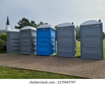 A Line Up Of Port A Potties Going Along A Walking Path. There Are Five Portable Bathrooms That Are Blue, Gray, And White.