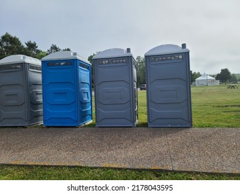 A Line Up Of Port A Potties Going Along A Walking Path. There Are Four Portable Bathrooms That Are Blue, Gray, And White.