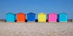 A Line Of Six Brightly Coloured Beach Huts, Each One Is A Different Colour In The Foreground Is A Pebble Beach And Behind Is A Bright Blue Sky, Seaford, East Sussex, England, United Kingdom, UK, 