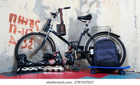 a linear composition of a bicycle, roller skates, a blue skateboard and a backpack standing on a tiled floor against an old concrete wall