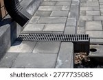 Linear drainage grate installed between concrete pavers, connected to downspout and catch basin, residential hardscape water management system, spring maintenance. Real photo