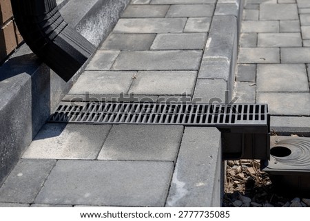 Linear drainage grate installed between concrete pavers, connected to downspout and catch basin, residential hardscape water management system, spring maintenance. Real photo
