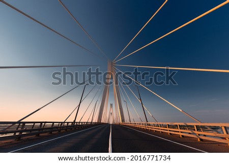 Linear perspective view of a white cable-stayed suspension bridge in the golden light of the rising sun shot from a ground level in horizontal (landscape) format