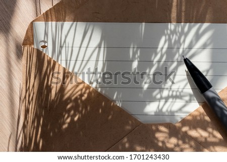 Lined sheet of paper (empty space for text) in craft envelope lies on the table, next to it is a gray mechanical pencil. Still life in the rays of the sunset or dawn sun, the shadow of plants
