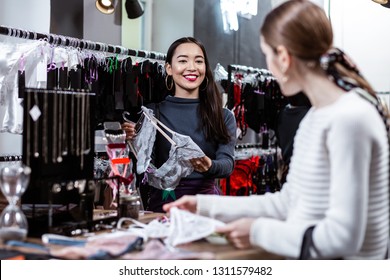 Lingerie collection. Dark-haired smiling asian lady wearing a dark-blue blouse discussing new lingerie collection with her friend