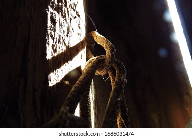 Links Of An Old, Rusty Chain Wrapped Around A Wooden Beam, Bright Light Shining Into The Heavily Shadowed Building Area Through A Slanted Or Diagonal Crack Between Two Boards On The Wall. 