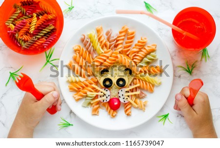 Lion pasta - fun food idea for kids lunch, animal shaped food art. Colorful fusilli vegetables pasta with sandwich like a cute lion head on white plate top view