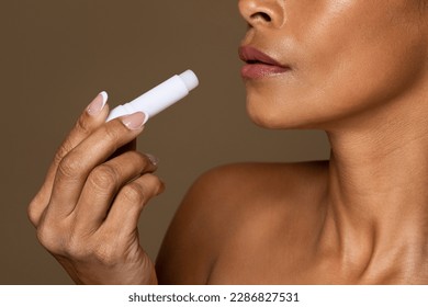 Lips care. Black middle aged woman applying lip balm, female using hygienic lipstick, lady with bare shoulders standing over brown studio background, closeup, copy space
