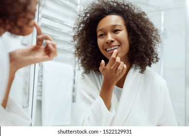 Lips Skin Care. Woman Applying Lip Balm Looking In Mirror At Bathroom. Portrait Of Beautiful African Girl Model With Beauty Face And Natural Makeup Applying Lip Product With Finger