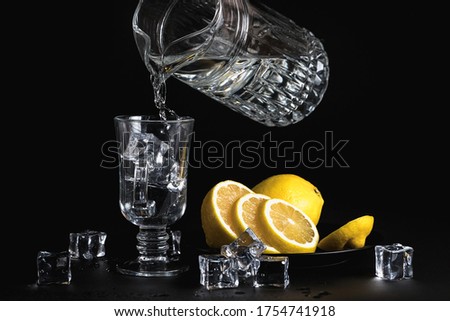Liquid from a glass jug pours into a stemmed glass. Still life on a black background. Refreshment. Sliced lemon. Ice cube.