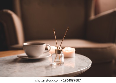 Liquid home fragrance with glass bottle and wooden sticks, cup of fresh tea staying on marble coffee table at home close up. Cozy atmosphere. 