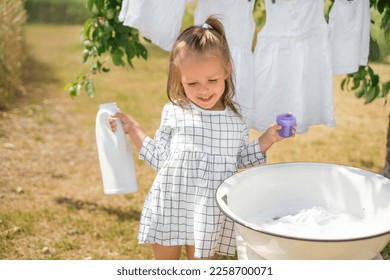 Liquid hypoallergenic organic washing powder for washing baby clothes. The child is holding a large white bottle of soap. Cute beautiful happy girl doing laundry - Shutterstock ID 2258700071