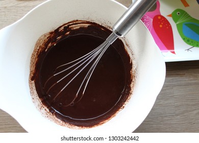 Liquid Melted Dark Chocolate With Whisk Inside A White Round Bowl. Top View, Overhead Closeup Shot With Wood Background