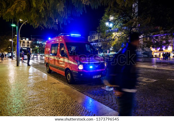 LISBON, PORTUGAL - CIRCA MARCH 2019: Long\
exposure of some people passing by an ambulance on duty with it\'s\
lights on, at night in\
\