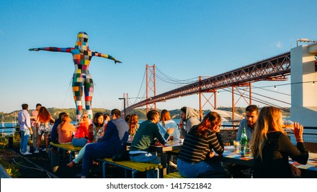 Lisbon, Portugal - June 5, 2019: View of 25th of April bridge with people sitting on the bar terrace at LX Factory in Alcantara during the sunset in Lisbon city, Portugal