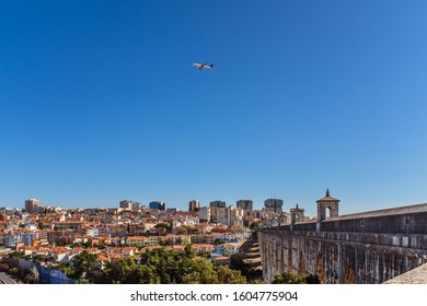 LISBON, PORTUGAL - OCTOBER 10, 2019: Plane Flying Over Águas Livres Aqueduct With Cityscape On A Background, Horizontal Crop.