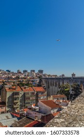 LISBON, PORTUGAL - OCTOBER 10, 2019: Plane Flying Over Águas Livres Aqueduct With Cityscape On A Background, Vertical Crop.