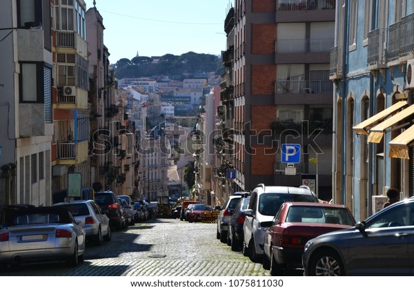 Lisbon street full of parked cars,\
street construction work ahead, hill view in the\
background