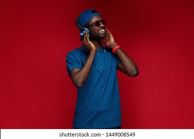 Listen Music. Smiling Black Man In Headphones Listening Sounds Portrait On Red Background. Happy Male Model In Stylish Sunglasses And Fashion Wear At Studio