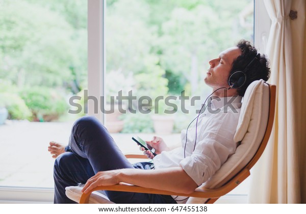 listening relaxing music\
at home, relaxed man in headphones sitting in deck chair in modern\
bright interior