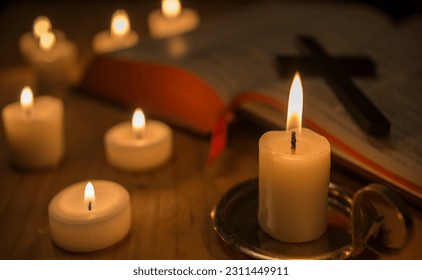  A lit candle in the foreground, lit candles in the background. Bible open and cross on the table
