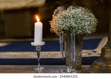 A lit candle in a glass holder beside a bouquet of baby's breath flowers in a vase, creating a warm and serene atmosphere. - Powered by Shutterstock