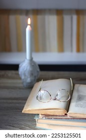 Lit candle, stack of books and reading glasses. Bookshelf in the background. Selective focus.