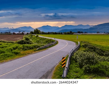 A lit rural road winding through green fields with mountain views during sunset in BC, Canada.