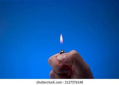 Lit small lighter with flame held between fingers by man. Close up studio shot, isolated on blue background.