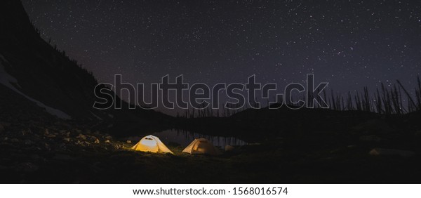 Lit tent at night in the\
Sawtooth Mountains of Idaho with stars in the sky above next to an\
alpine lake