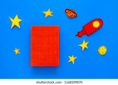 Literature For Children. Fantactic, Fiction Story. Book With Blank Cover Near Cutout Of Rocket, Stars, Moon On Blue Background Top View Mockup