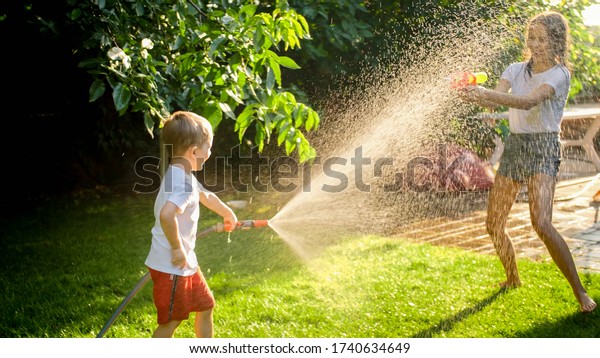 Litle boy having water fight on the backyard with\
his sister.