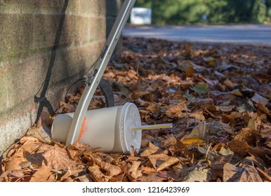 A Litter Picker Using A Tool Retrieves A Discarded Soda Cup With Straw That Lies Amongst Fallen Leaves At The Base Of A Brick Wall In The Autumn