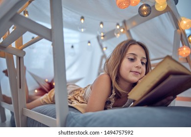 Little 10 Year Old Girl Reading Classic Book Under Her Home-made Tent Inside The Living Room.