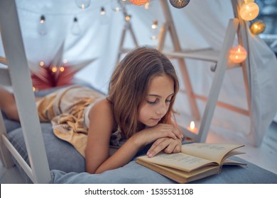 Little 10 year old girl reading classic book under her home-made tent inside the living room.