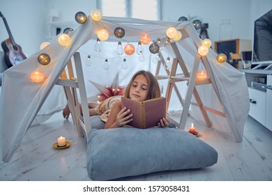 Little 10 Year Old Girl Reading Classic Book Under Her Home-made Tent Inside The Living Room.