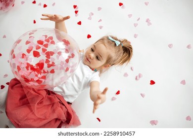 Little 2 years old girl playing with balloon with heart shape papers inside it and around, lying on white background. Valentines Day concept