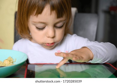 Little 3 year old girl having lunch at home while playing on a tablet.