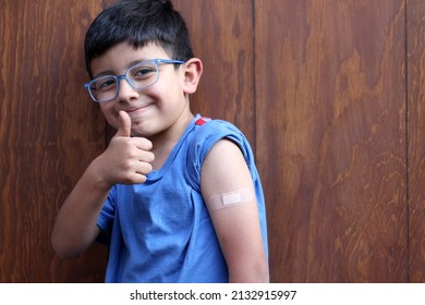 Little 6-year-old Latino boy with glasses and a blue shirt shows his arm with a bandage because he has just been vaccinated against Covid-19 in the new normality due to the Coronavirus pandemic
