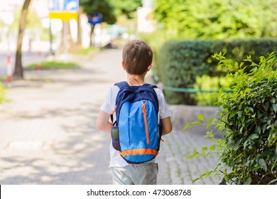 Little 7 Years Schoolboy Going To School. Dressed In White T Shirt And Shorts. Blue Backpack