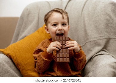 Little Adorable Boy Sitting On The Couch At Home And Eating Chocolate Bar. Child And Sweets, Sugar Confectionery. Kid Enjoy A Delicious Dessert. Preschool Child With Casual Clothing. Positive Emotion.