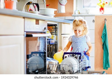 Little Adorable Cute Toddler Girl Helping To Unload Dishwasher. Funny Happy Child Standing In The Kitchen, Holding Dishes And Putting A Bowl On Head. Healthy Kid At Home. Gorgeous Helper Having Fun