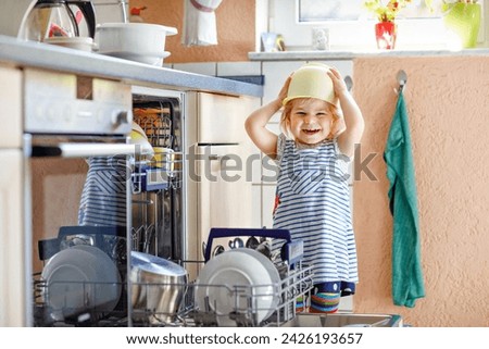 Little adorable cute toddler girl helping to unload dishwasher. Funny happy child standing in the kitchen, holding dishes and putting a bowl on head. Healthy kid at home. Gorgeous helper having fun