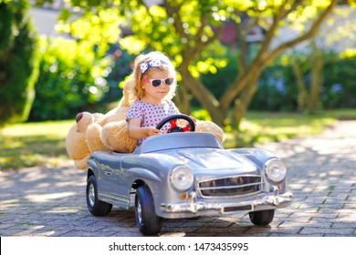 Little adorable toddler girl driving big vintage toy car and having fun with playingoutdoors. Gorgeous happy healthy child enjoying warm summer day. Smiling stunning kid playing in domestic garden,