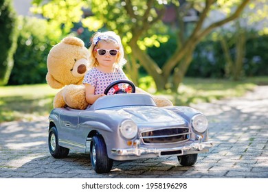 Little adorable toddler girl driving big vintage toy car and having fun with playing with plush toy bear, outdoors. Gorgeous happy healthy child enjoying warm summer day. Smiling stunning kid in gaden