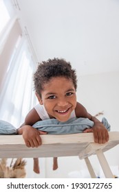 Little African American Boy Has Fun, Lies On White Wooden Chair, Flounders With His Feet. Minimalist Interior, Pampas Grass, Driftwood On The Wall, Airy Doors On The Windows. Scandinavian Style.