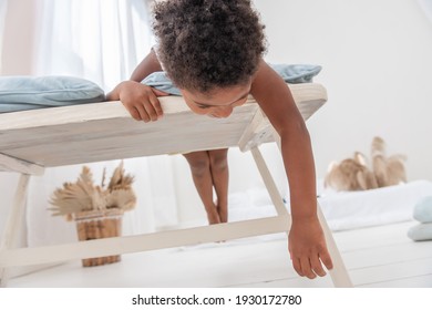 Little African American Boy Has Fun, Lies On White Wooden Chair, Flounders With His Feet. Minimalist Interior, Pampas Grass, Driftwood On The Wall, Airy Doors On The Windows. Scandinavian Style.