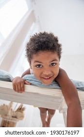 Little African American Boy Has Fun, Lies On White Wooden Chair, Flounders With His Feet. Minimalist Interior, Pampas Grass, Driftwood On The Wall, Airy Doors On The Windows. Scandinavian Style.