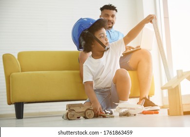 A Little African American Boy Wear Engineering Helmet Pretend To Be Engineer Play With Wooden Toys On The Floor While Dad Sitting And Looking At His Son On Background