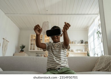 Little african american boy wearing 3d virtual reality glasses sits on couch at home. Online world simulation and play game, racing drives car. Teenager gesturing hands in air watches in vr goggles - Shutterstock ID 2314950701